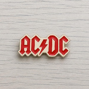 Puede incluir: Un pin con el logotipo de AC/DC en rojo y blanco. El pin presenta el nombre de la banda en una fuente audaz y estilizada con un rayo en el centro. El pin tiene un borde dorado y está sobre un fondo de vetas de madera clara.
