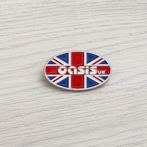 Może przedstawiać: Owalna przypinka z motywem Union Jack w kolorach czerwonym, białym i niebieskim. Słowo "Oasis" jest w białych blokowych literach, z "UK" poniżej. Przypinka ma srebrną obwódkę i jest umieszczona na jasnym tle słojów drewna.