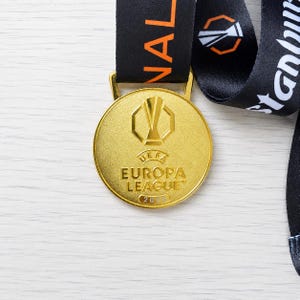 以下が含まれることがあります： 表面にUEFAヨーロッパリーグのロゴと「UEFA EUROPA LEAGUE 2016」の文字が刻印された、金色の円形メダル。メダルは黒い布製のリボンに取り付けられており、上部にはオレンジと白の文字のディテールが見えます。