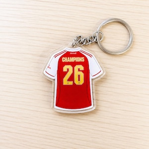 Peut inclure: Un porte-clés rouge et blanc en forme de maillot de football. Le numéro 26 en jaune est affiché au dos, avec le mot "CHAMPIONS" au-dessus. Le porte-clés est attaché à un anneau métallique argenté.