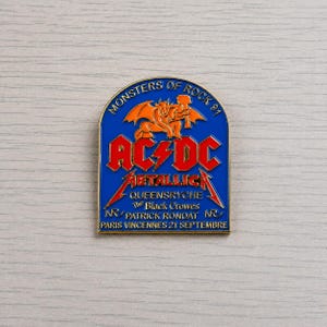 Puede incluir: Pin metálico azul y dorado con el texto "MONSTERS OF ROCK 91" y los nombres de las bandas AC/DC y Metallica. El pin también enumera Queensryche, The Black Crowes y Patrick Rondat, con "PARIS VINCENNES 21 SEPTEMBRE" en la parte inferior.