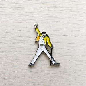 Könnte beinhalten: Emaille-Anstecknadel mit einer stilisierten Darstellung eines Künstlers in einem weißen Overall, einer gelben Jacke und einem schwarzen Schnurrbart, der einen Mikrofonständer mit erhobenem Arm hält. Die Nadel hat eine schwarze Umrandung.