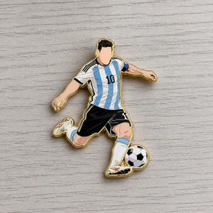 May include: A gold-colored pin depicting a soccer player in a light blue and white striped jersey with the number 10, black shorts, and white socks. The player is kicking a black and white soccer ball.