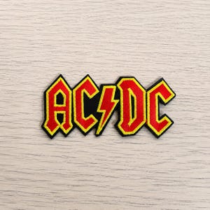 May include: An embroidered fabric patch featuring the AC/DC logo in red with a bright yellow border. The design includes the letters A, C, D, and C with a central lightning bolt symbol, all outlined in black thread against a light wood background.
