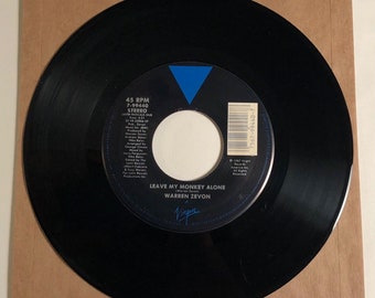 May include: A black vinyl record with a blue triangle and white label. The label has the text "45 RPM", "7-99440", "STEREO", "LATIN RASCAIS DUB", "LEAVE MY MONKEY ALONE", "WARREN ZEVON", and "Virgin".