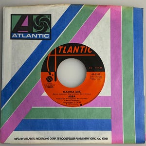 May include: A 45 RPM vinyl record by ABBA titled Mamma Mia, featuring an orange center label with black text. The record is housed in a square paper sleeve with a white background and large, stylized letter A graphics in green and purple.