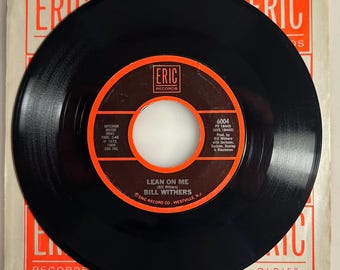 May include: A black vinyl record with an orange and white label featuring the song title "LEAN ON ME" by Bill Withers. The record is in its original paper sleeve, with "ERIC RECORDS" printed on it.