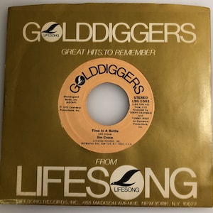 Puede incluir: Una funda de disco de vinilo de 45 RPM vintage con un diseño dorado y blanco. La etiqueta del disco es de color beige con texto negro, con la canción "Time In A Bottle" de Jim Croce. La funda dice "Golddiggers" y "Lifesong Records."