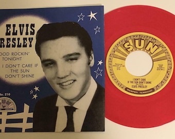 Può includere: Un disco in vinile rosso con un'etichetta gialla che mostra il testo "SUN RECORDS" e "ELVIS PRESLEY" con il titolo della canzone "I DON'T CARE IF THE SUN DON'T SHINE". Il disco è accanto a una foto in bianco e nero di Elvis Presley su uno sfondo blu con il testo "ELVIS PRESLEY" e "GOOD ROCKIN' TONIGHT" e "I DON'T CARE IF THE SUN DON'T SHINE".