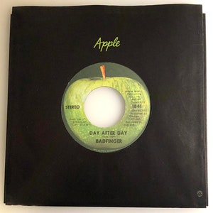 May include: A black and white 45 RPM vinyl record with a green apple on the cover. The record is titled "Day After Day" by Badfinger. The record label is Apple Records.