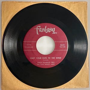 May include: A black vinyl record with a red label featuring the text "Fantasy" and "CAST YOUR FATE TO THE WIND" by Vince Guaraldi Trio. The record is in a tan paper sleeve.