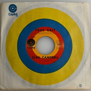 May include: A yellow and blue vinyl record with the text "TRUE GRIT" and "GLEN CAMPBELL" on it. The record is labeled "Capitol" and has the song title "True Grit" by "Don Black-Elmer Bernstein" from the Paramount picture "True Grit".