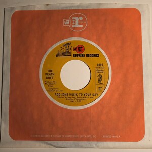 May include: A yellow and black vinyl record with the text "The Beach Boys" and "Add Some Music To Your Day" on it. The record is on a red label with the text "Reprise Records" and "0894 (N18242)".