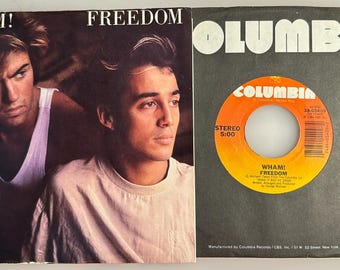 May include: A 7-inch vinyl record sleeve and record for Wham! Freedom. The sleeve features a photograph of two individuals, one in a white tank top and one in a white t-shirt. The orange record label displays the Columbia logo and text.