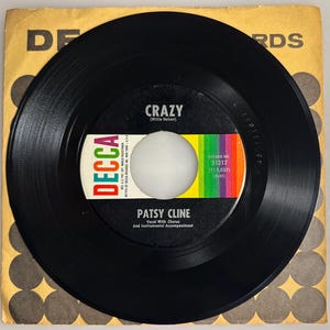 May include: A vintage 45 RPM vinyl record of Patsy Cline's song "Crazy" on the Decca Records label. The record is black with a colorful label featuring the song title, artist, and record information. The record is in its original yellow and brown sleeve.