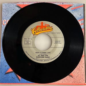 May include: A black vinyl record with a white label featuring the text "Keep It Comin' Love" by KC and The Sunshine Band. The record is on a colorful sleeve with red, blue, and white starburst designs. The label also includes the Collectables logo.