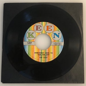 以下が含まれることがあります： 「EE K N」という文字と、サム・クックの曲「Everybody Likes to Cha Cha Cha」のタイトルが書かれたカラフルなラベルが付いた黒いビニールレコード。ラベルには、作曲者のバーバラ・キャンベルとレコードレーベルのKeen Productionsも記載されている。