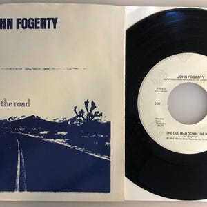 May include: A 45 RPM vinyl record by John Fogerty. The record is black with a white label. The label features the title "The Old Man Down The Road" and the artist's name "John Fogerty".