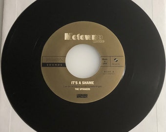 May include: A black vinyl record with a gold label. The label features the text "Motown & more", "Renation Sounds", "It's a Shame" by "The Spinners", and "Side A" with the song's catalog number. The record is made in the USA.