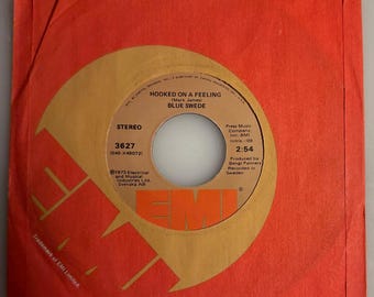 May include: A vintage 45 RPM record by Blue Swede, titled "Hooked on a Feeling." The record is in a red sleeve with a gold and orange label. The label includes the song title, artist, and other details.