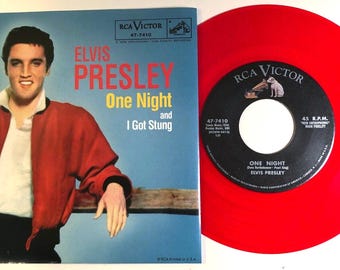 Può includere: Un disco in vinile rosso da 45 giri con un'etichetta nera. L'etichetta presenta un'immagine di un cane e il testo "RCA VICTOR" e "One Night" di "Elvis Presley".