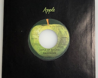 May include: A green apple record label with the text "Apple" in a cursive font. The label features a green apple with a leaf and the text "Apple of My Eye" by Badfinger. The record label is from the LP "HASS" W-3411. The label also includes the text "Apple Music Publishing Co. Inc. ASCAP 1864" and "Produced by Chris Thomas and Badfinger. Recorded in England."