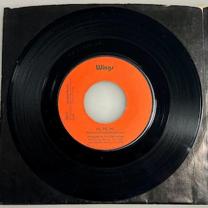 May include: A black vinyl record with an orange label featuring the text "Wings" and "Hi, Hi, Hi" by Paul and Linda McCartney. The record is in a black paper sleeve. The label also includes the text "An Apple Record".
