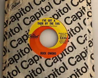 May include: A black and white 45 RPM vinyl record with a yellow and orange label. The record is titled "I've Got a Tiger By The Tail" by Buck Owens. The label also includes the songwriters' names, the record label, the song's BMI number, and the record number.