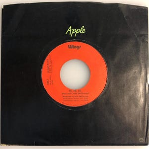 May include: A vintage vinyl record sleeve featuring the song "Hi, Hi, Hi" by Wings. The record label is orange with black text, set against a black sleeve. The Apple Records logo and Wings logo are visible.