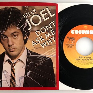 Può includere: Un disco in vinile di Billy Joel con la canzone "Don't Ask Me Why" sull'etichetta. Il disco è nero con un'etichetta gialla. La copertina presenta una foto di Billy Joel con del vetro rotto davanti a lui.