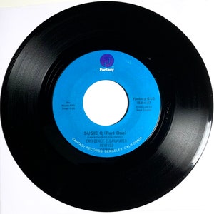 May include: A black vinyl record with a blue label. The label has a white circle in the center and the text "Fantasy 616 (Side A)" and "Susie Q (Part One)" printed on it. The record is from the band Creedence Clearwater Revival.