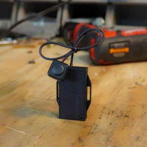 Puede incluir: Un componente electrónico negro con un botón y una carcasa rectangular. El botón redondo está conectado a la carcasa por un cable negro. La carcasa tiene una superficie texturizada y dos aberturas rectangulares.