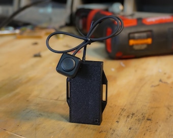 Puede incluir: Un componente electrónico negro con un botón y una carcasa rectangular. El botón redondo está conectado a la carcasa por un cable negro. La carcasa tiene una superficie texturizada y dos aberturas rectangulares.