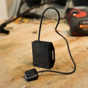 May include: Black rectangular device with a textured surface, a handle-like cutout, and a connected button. A black cable connects the device to a small square button. The device is on a wooden surface.