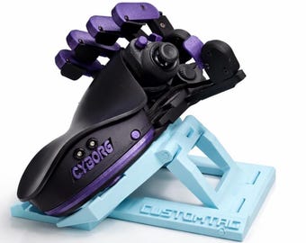 May include: A black and purple mechanical prosthetic hand with the word CYBORG printed on the side, mounted on a light blue plastic stand with the word CUSTOMTAG embossed on the base. The device features articulated fingers and a central joystick.