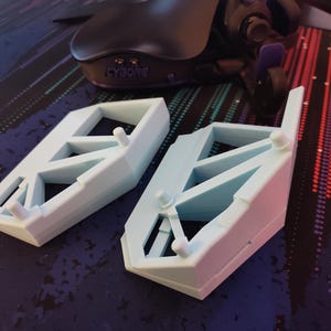 May include: Two light blue, geometric-shaped objects with a rectangular base and a series of internal supports. The objects have small cylindrical protrusions. A black gaming mouse is visible in the background.