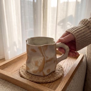 Könnte beinhalten: Eine beige Keramiktasse mit einem einzigartigen, strukturierten Design und einem gebogenen Griff. Die Tasse steht auf einem runden, gewebten Untersetzer, der auf einem Holztablett platziert ist. Das Innere der Tasse ist heller, und die Außenseite hat braune Marmorierungen.