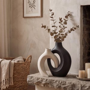Może przedstawiać: Dwa nowoczesne ceramiczne wazony, jeden kremowy i jeden czarny, stoją na kamiennej powierzchni. Czarny wazon zawiera suszone gałązki eukaliptusa. Pleciony kosz z kocem i oprawiony botaniczny nadruk znajdują się w tle.
