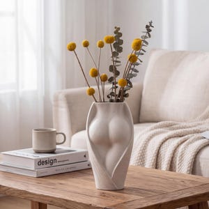 May include: A white ceramic vase with a unique form, resembling a torso, holds yellow dried flowers and eucalyptus. A beige mug and books are on a wooden table. The vase is a decorative home accent.