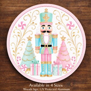 May include: A round, pink-edged sign featuring a watercolor illustration of a nutcracker soldier, Christmas trees, and gifts. The nutcracker is in blue and pink with gold accents. The sign has a white background with gold flourishes and candy canes.