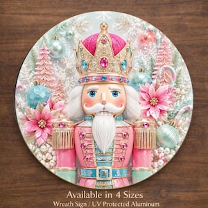 May include: A circular aluminum wreath sign featuring a nutcracker wearing a pink and gold crown with blue gems. The nutcracker has a white beard and pink uniform with gold accents. Background includes pink trees, blue ornaments, and poinsettias.