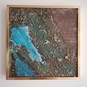 Op de afbeelding: Abstract textuurschilderij in een gouden lijst. Het kunstwerk toont een mix van kleuren, waaronder blauw, groen en bruin, met een gebarsten, gelaagd effect. Het schilderij is vierkant en gemonteerd in een goudkleurige lijst.