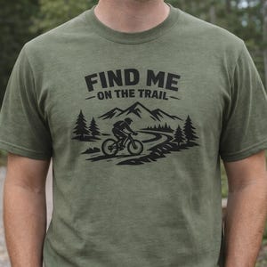 Könnte beinhalten: Olivgrünes T-Shirt mit dem Text "FIND ME - ON THE TRAIL" über einer schwarzen Grafik eines Mountainbikers auf einem kurvenreichen Pfad. Das Design umfasst Berge und Bäume und schafft ein Design zum Thema Natur.