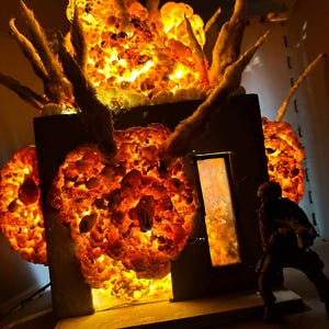 May include: A diorama featuring a dark gray cube structure engulfed in bright orange and yellow illuminated explosions, with textured debris plumes extending upward. A small dark soldier figurine stands near a rectangular doorway.