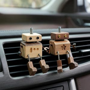 May include: Two wooden robot air fresheners with light and dark wood tones. Each robot has a square head and body, with jointed arms and legs. One robot has a star cutout, the other has three small holes. They are attached to a car vent.