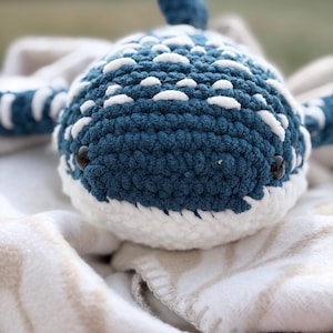 May include: A crocheted whale shark plush toy featuring a deep blue body with white stitched patterns, a white underbelly, and two small black plastic eyes. The plush has a rounded head, side fins, and a dorsal fin visible on top.