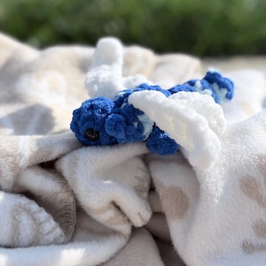 May include: A small, handcrafted crochet dragonfly toy featuring a textured blue and white body with large, soft white wings. The dragonfly has a rounded head with a single visible dark eye and a segmented tail, constructed from plush yarn.