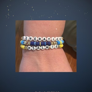 May include: Three stacked bracelets with white letter beads spelling "GENERATION" and "PSALM 78". The bracelets feature yellow, blue, and navy beads with gold accents. The background is a dark blue with small white stars.