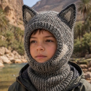 May include: A gray and white knit balaclava with pointed ears. The balaclava covers the head and neck, with an opening for the face. The hat is made of a textured yarn and is worn by a person wearing a green jacket.