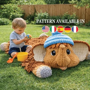 May include: A crocheted elephant-shaped baby blanket in brown, tan, and blue. The blanket has large ears and a trunk, with a blue hat on the elephant's head. A young child is playing with toys on the grass near the blanket. The text "PATTERN AVAILABLE IN" is visible.